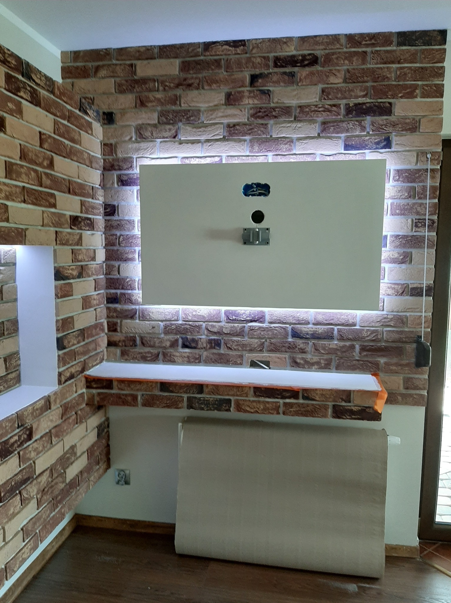 Ściana z cegły dekoracyjnej z podświetleniem LED, półka i przygotowane miejsce na telewizor. Widoczne gniazdka elektryczne i fragment grzejnika.