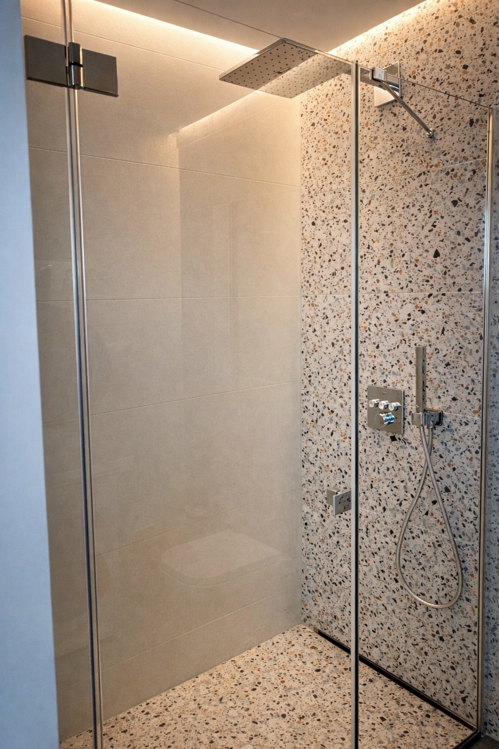 Nowoczesna kabina prysznicowa z deszczownicą, wykończona płytkami w kolorze terazzo i jasnymi płytkami ceramicznymi. Szklane drzwi z chromowanymi zawiasami.