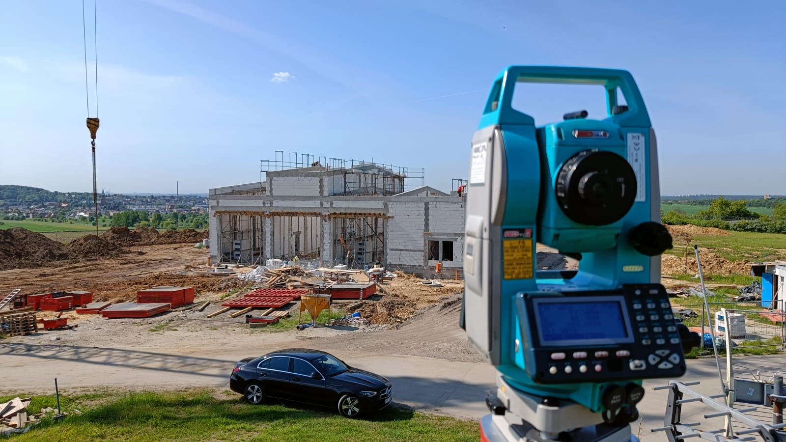Niwelator elektroniczny na budowie domu jednorodzinnego w stanie surowym otwartym, z widocznym dźwigiem i czarnym samochodem osobowym w tle.