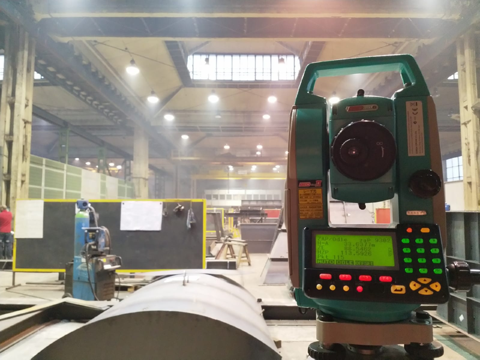 Zielony teodolit elektroniczny na tle hali produkcyjnej z widocznymi elementami konstrukcyjnymi i oświetleniem. Ekran urządzenia wyświetla dane pomiarowe.