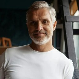 Dominik Kul - Portret uśmiechniętego mężczyzny z siwą brodą w białej koszulce, opierającego się o czarną konstrukcję. Ujęcie z bliska, naturalne światło.