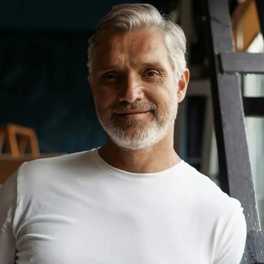 Portret uśmiechniętego mężczyzny z siwą brodą w białej koszulce, opierającego się o czarną konstrukcję. Ujęcie z bliska, naturalne światło.