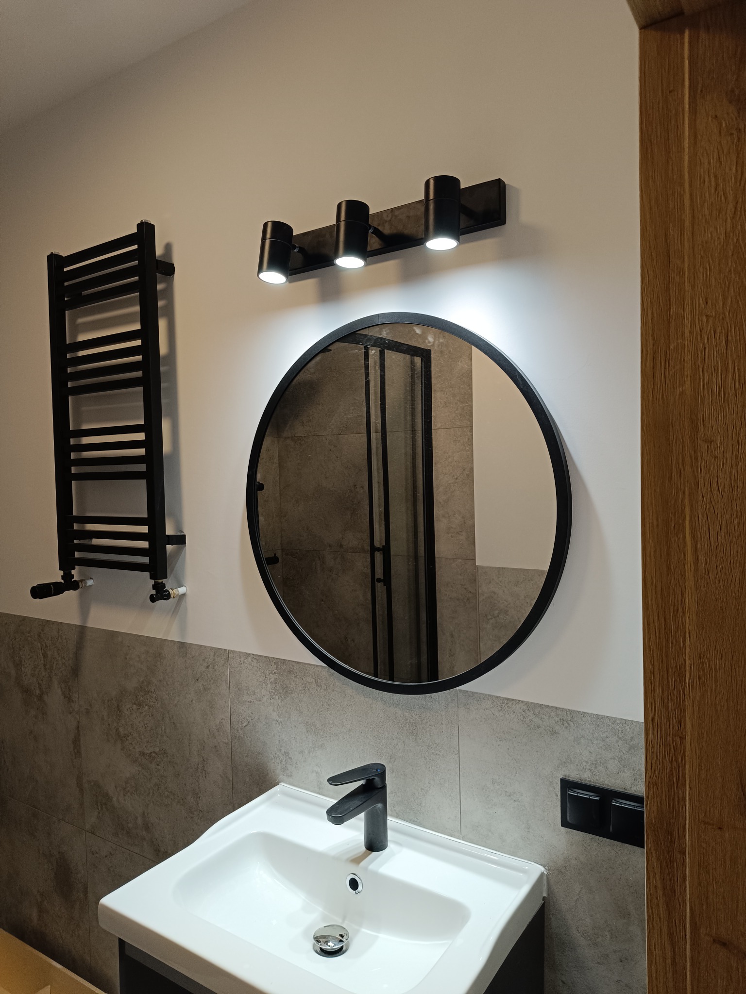 Nowoczesna łazienka z okrągłym lustrem, czarną armaturą i grzejnikiem drabinkowym. Szare płytki kontrastują z bielą ścian i ceramiki. Minimalistyczny design.