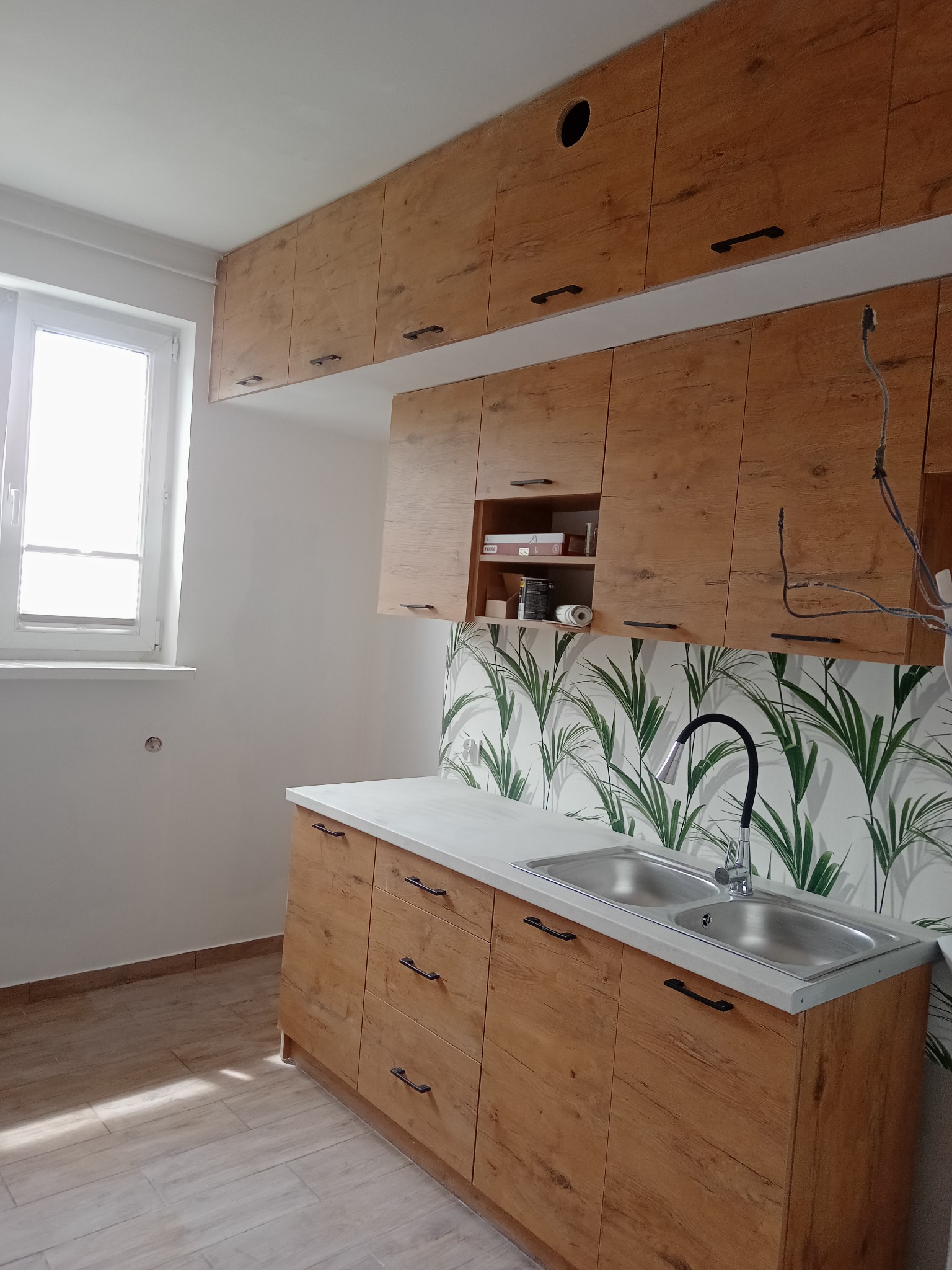 Jasna kuchnia z drewnianymi szafkami i blatem, wzorzysta ściana nad zlewem, okno z roletą. Widoczne niedokończone podłączenia elektryczne.