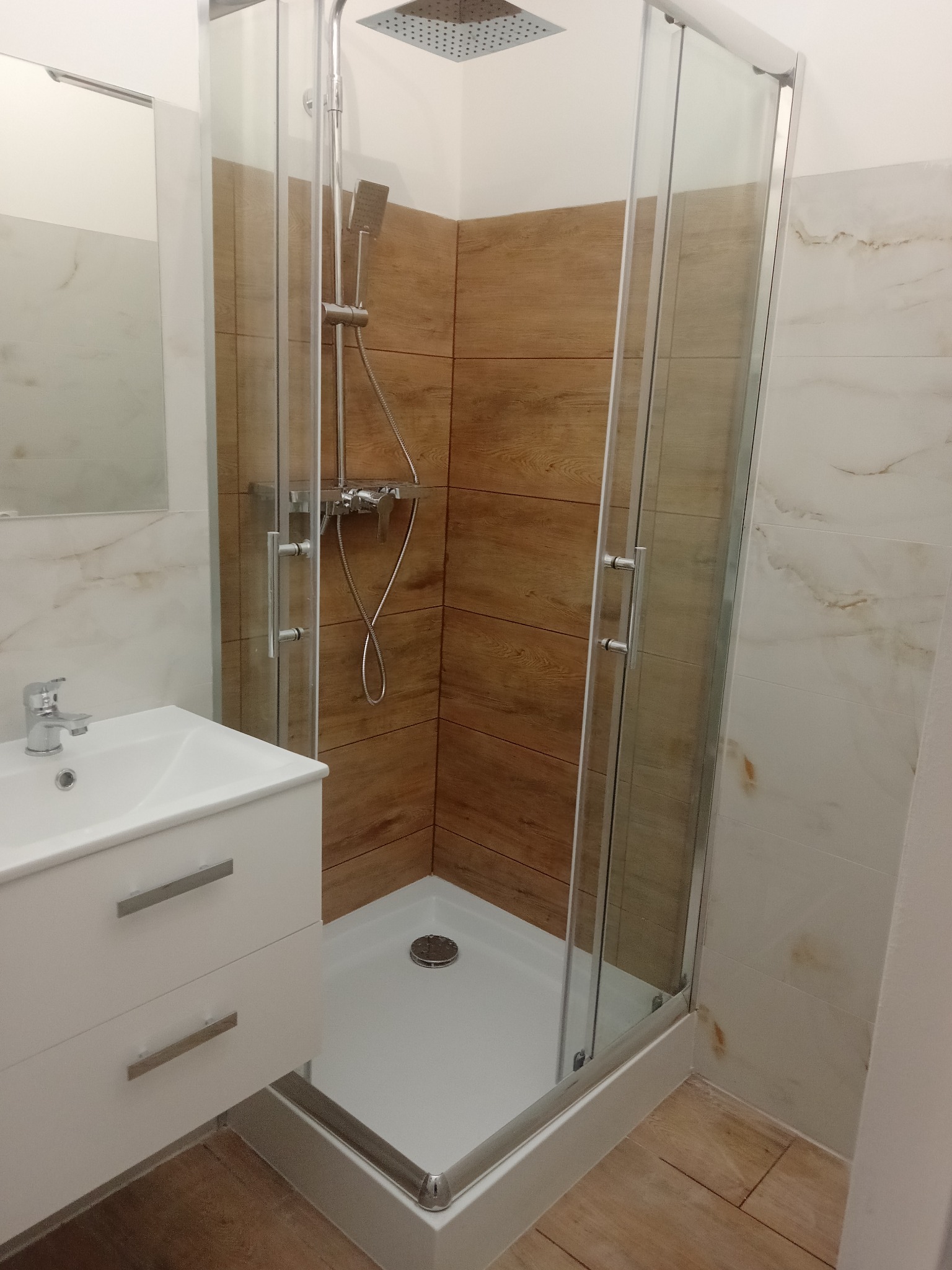 Nowoczesna kabina prysznicowa z deszczownicą i szklanymi drzwiami, obok biała umywalka z szafką. Ściany wyłożone płytkami imitującymi drewno i marmur.