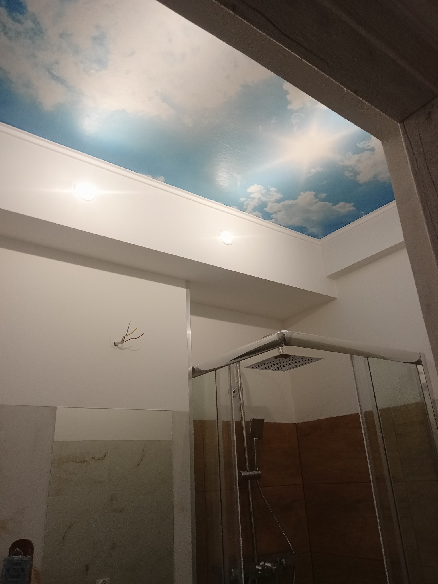 Wykończona łazienka z kabiną prysznicową i sufitem z motywem nieba. Widoczne oświetlenie punktowe i fragment lustra. Nietypowe wykończenie sufitu.