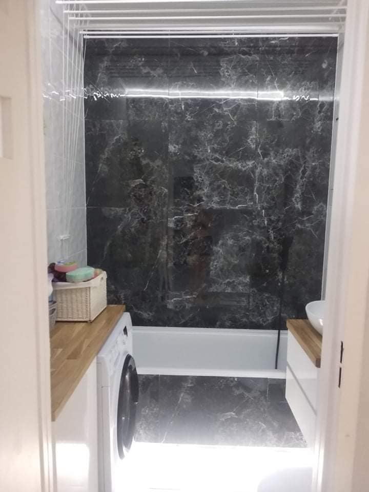 Wykończona łazienka z czarnymi płytkami imitującymi marmur na ścianach i podłodze. Widoczna pralka pod blatem z jasnego drewna i biała wanna.