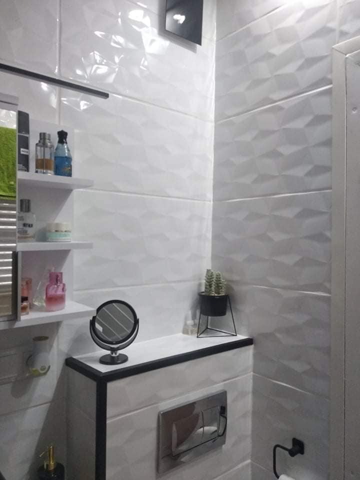 Biała łazienka z trójwymiarowymi płytkami, czarnymi akcentami i minimalistycznymi półkami. Na blacie małe lusterko i kaktus w czarnej doniczce.