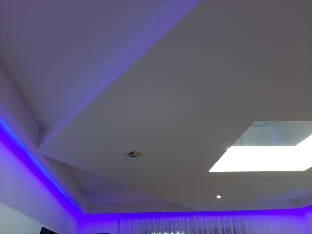 Nowoczesny sufit podwieszany z oświetleniem LED w kolorze fioletowym, widok z dołu. Geometryczne kształty i białe wykończenie. Centralnie umieszczone okno dachowe.