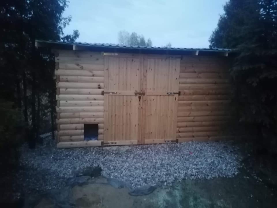 Drewniana konstrukcja z dwuskrzydłowymi drzwiami i małym otworem, otoczona kamieniami. Budowla z bali w otoczeniu zieleni, widoczna w słabym oświetleniu.