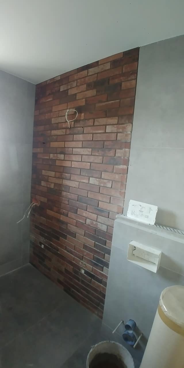 Ściana w łazience wyłożona cegłą dekoracyjną w odcieniach brązu i szarości, z widocznymi przewodami elektrycznymi. Obok fragment ściany wykończonej szarą płytą.