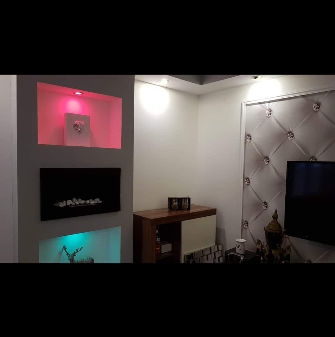 Nowoczesny salon z podświetlanymi wnękami w ścianie, dekoracyjnym panelem tapicerowanym i komodą z elementami drewna. Oświetlenie LED w różnych kolorach.