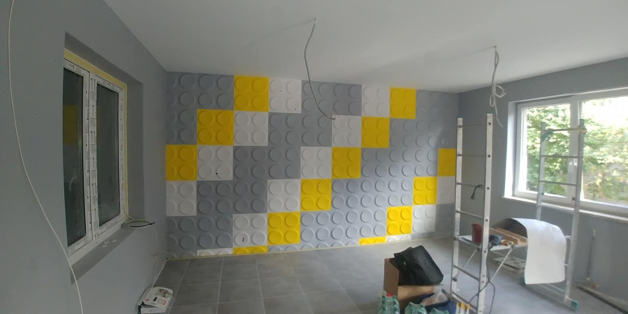 Pokój po remoncie: ściana z panelami 3D w szarościach, bieli i żółci, ułożonymi w szachownicę. Widoczne okno, drabina i podłoga z szarych płytek.
