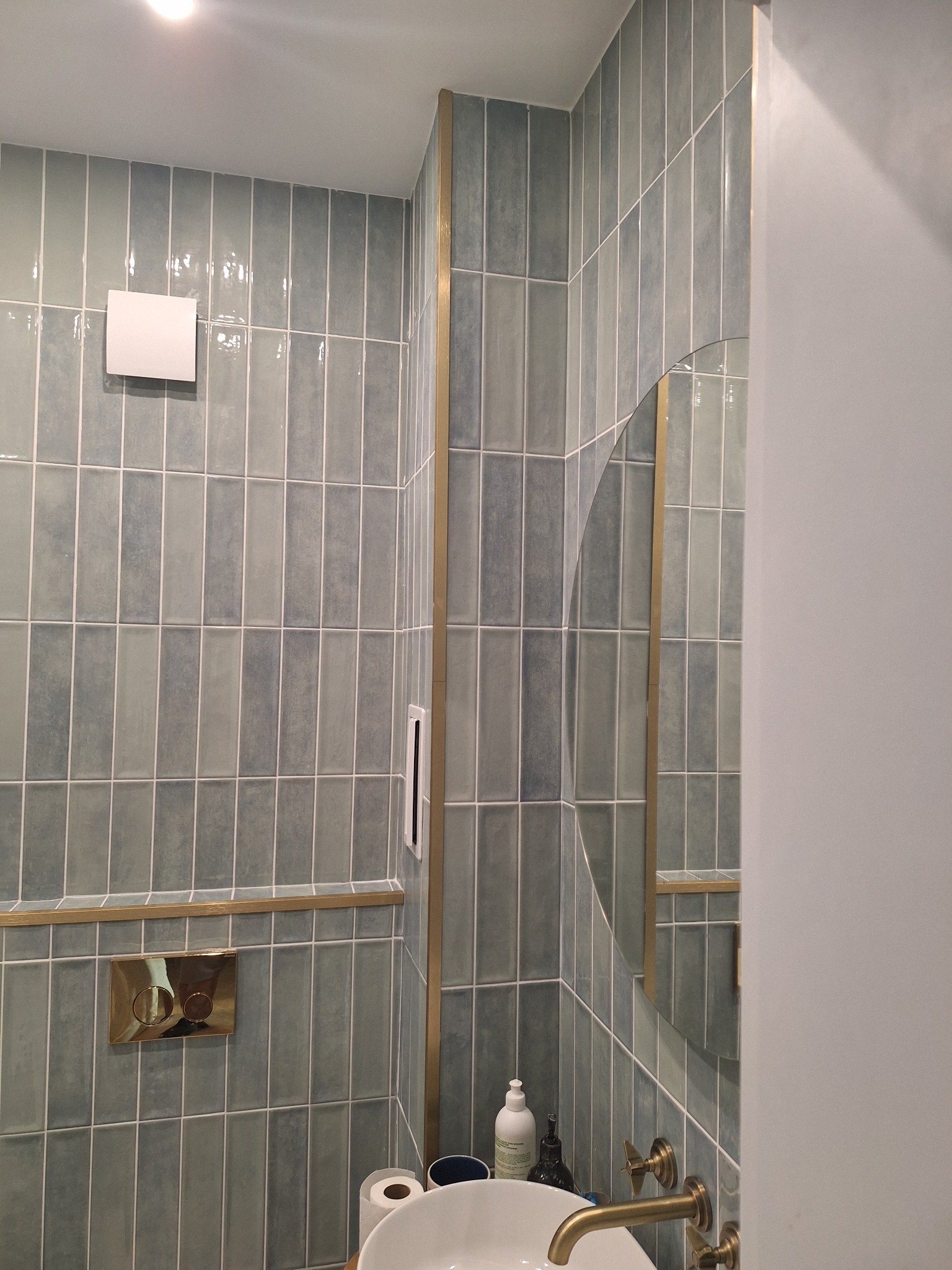 Błękitne, pionowe płytki w łazience z złotymi akcentami. Okrągłe lustro, biała umywalka i złota bateria. Widoczny fragment ściany i sufitu z oświetleniem.