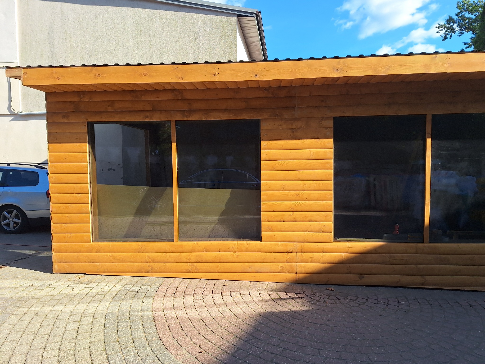 Drewniana konstrukcja z trzema oknami na brukowanym podłożu, prawdopodobnie altana lub mały budynek gospodarczy. Widoczny fragment samochodu i budynku w tle.