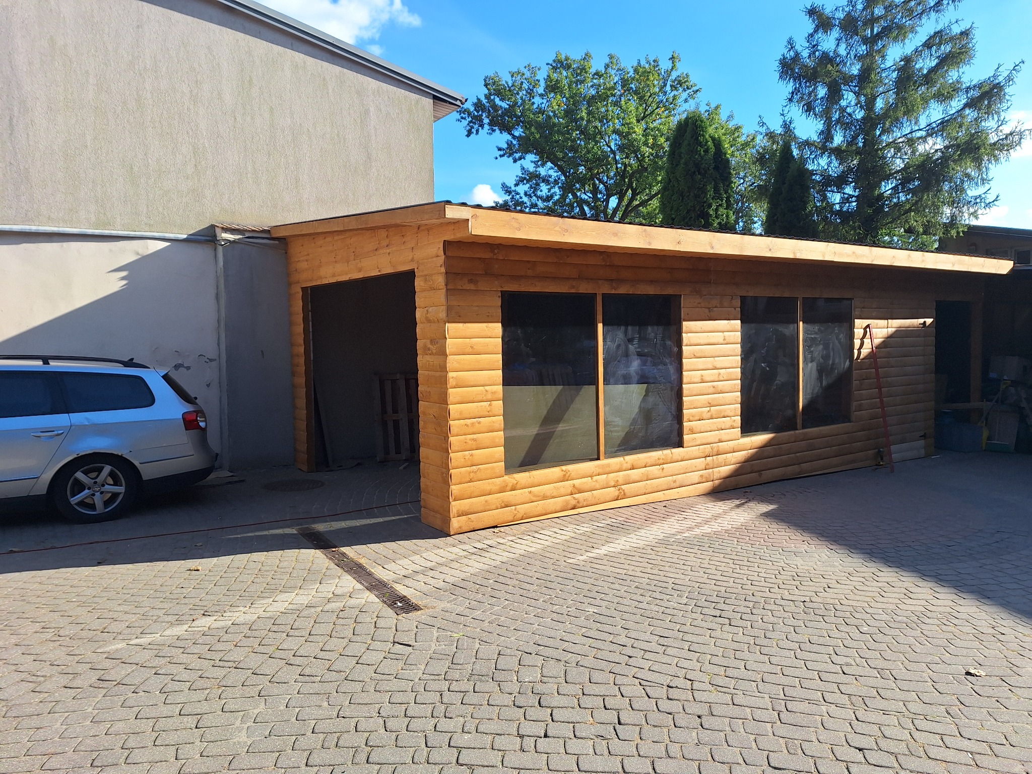 Drewniana pergola z oknami, przylegająca do budynku, z zaparkowanym samochodem obok. Kostka brukowa na podłożu, błękitne niebo w tle.