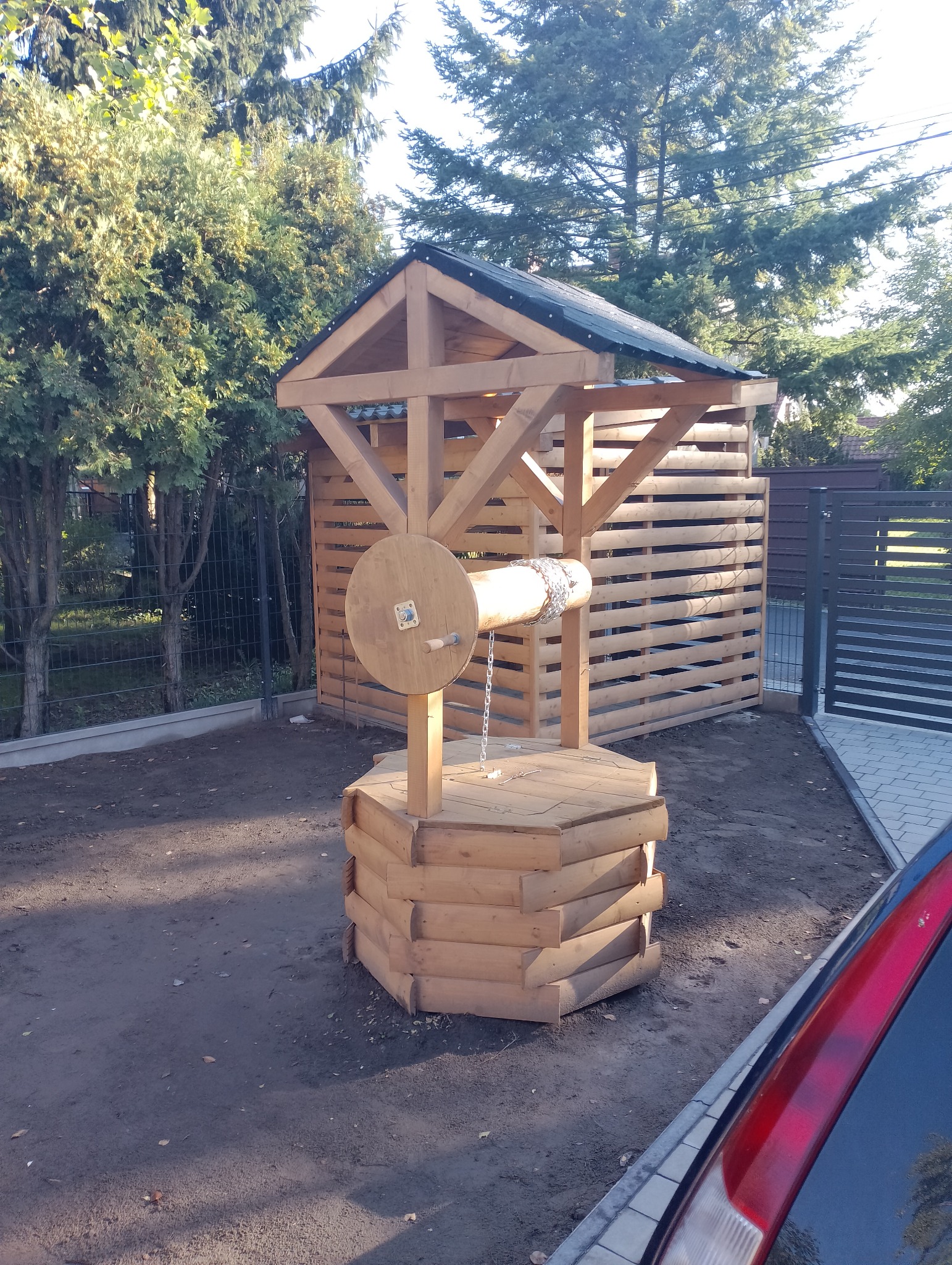 Drewniana studnia ogrodowa z zadaszeniem, wykonana z jasnego drewna, z widoczną konstrukcją i kołowrotem. W tle ogrodzenie z drewnianych listew i zieleń drzew.