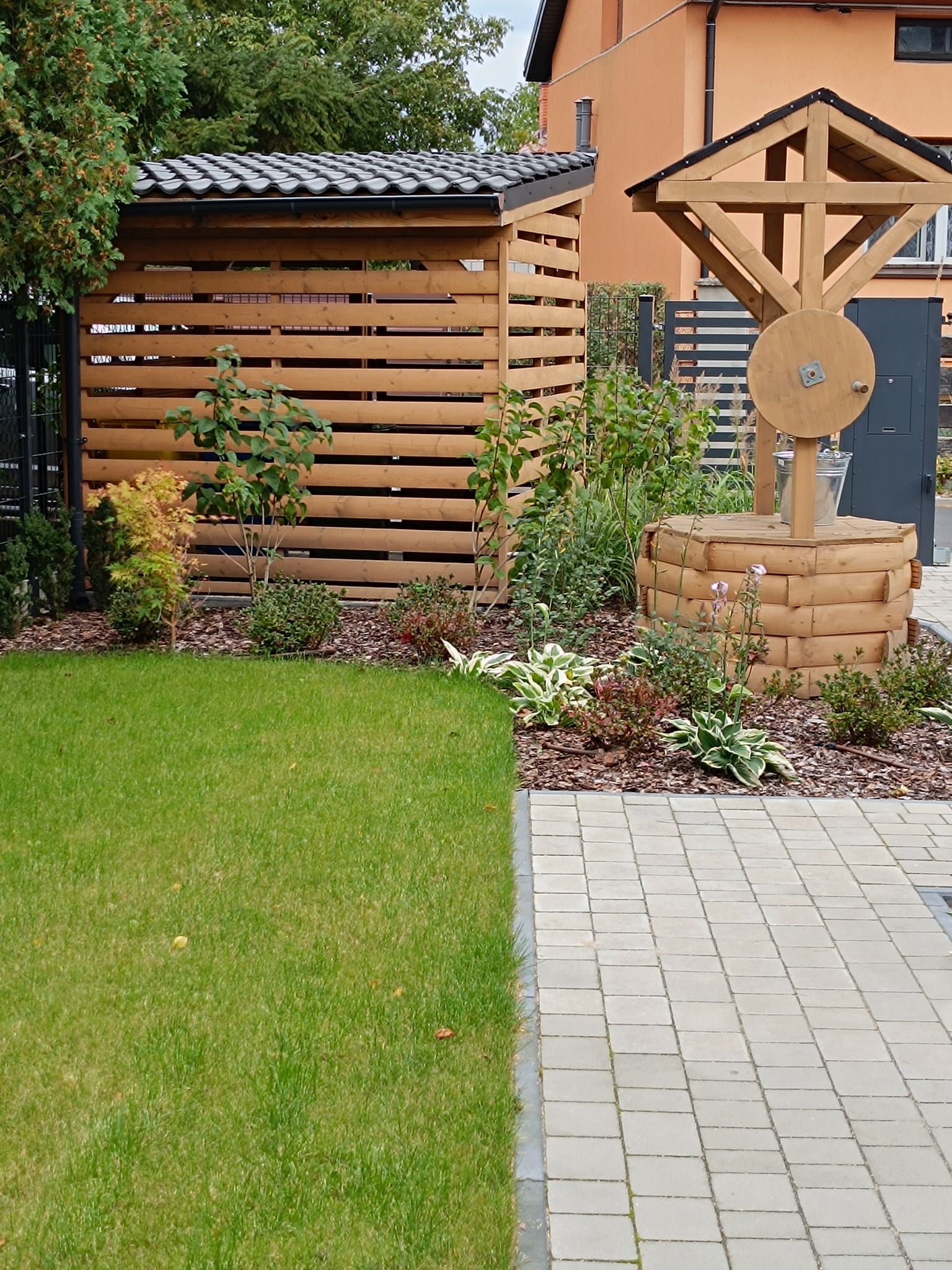 Drewniana studnia z wiaderkiem i zadaszona konstrukcja z poziomych desek w ogrodzie z kostką brukową i trawnikiem. Elementy małej architektury ogrodowej.