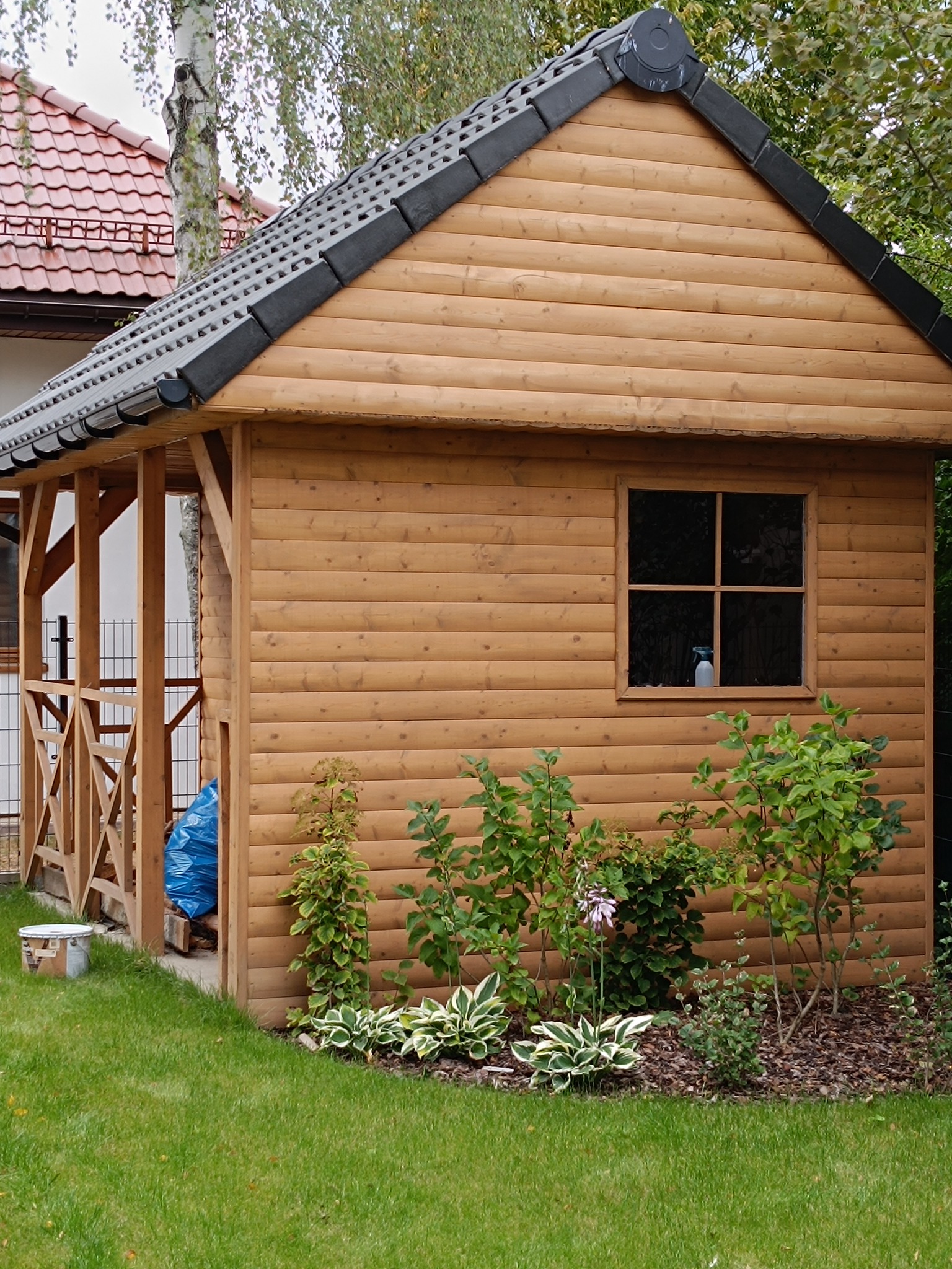 Drewniana altana z oknem i czarnym dachem, otoczona zielenią. Widoczny fragment domu z czerwoną dachówką w tle. Na trawie stoi wiadro.