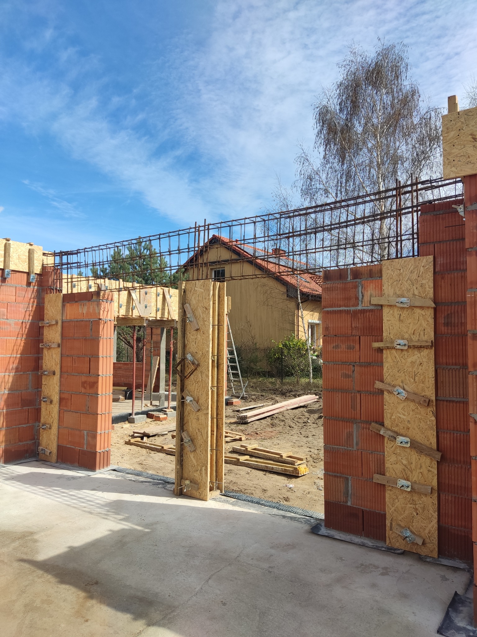 Wznoszenie ścian z czerwonej cegły z drewnianym szalunkiem i zbrojeniem. Widoczny fundament, teren budowy i dom w tle pod błękitnym niebem.