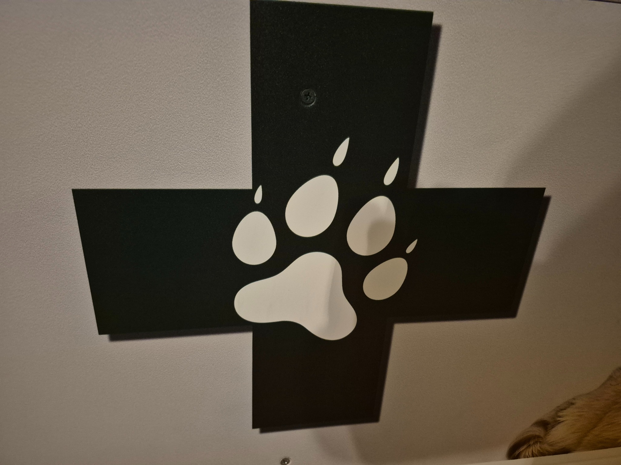 Czarny krzyż na ścianie z białą łapą zwierzęcia, przymocowany śrubami. Dekoracja wnętrza z motywem zwierzęcym, prawdopodobnie weterynaryjnym.