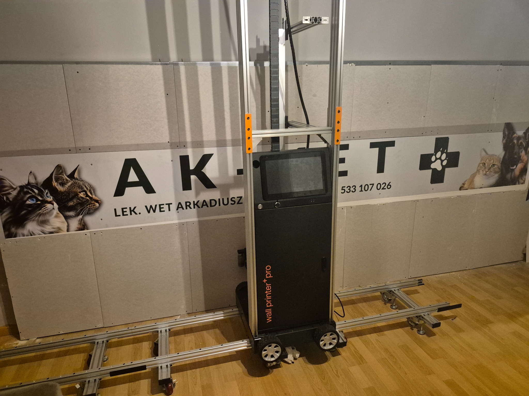 Ściana z płyt gipsowo-kartonowych przed drukiem pionowym drukarką 'wall printer pro', widoczne szyny i urządzenie drukujące. W tle reklama gabinetu weterynaryjnego.