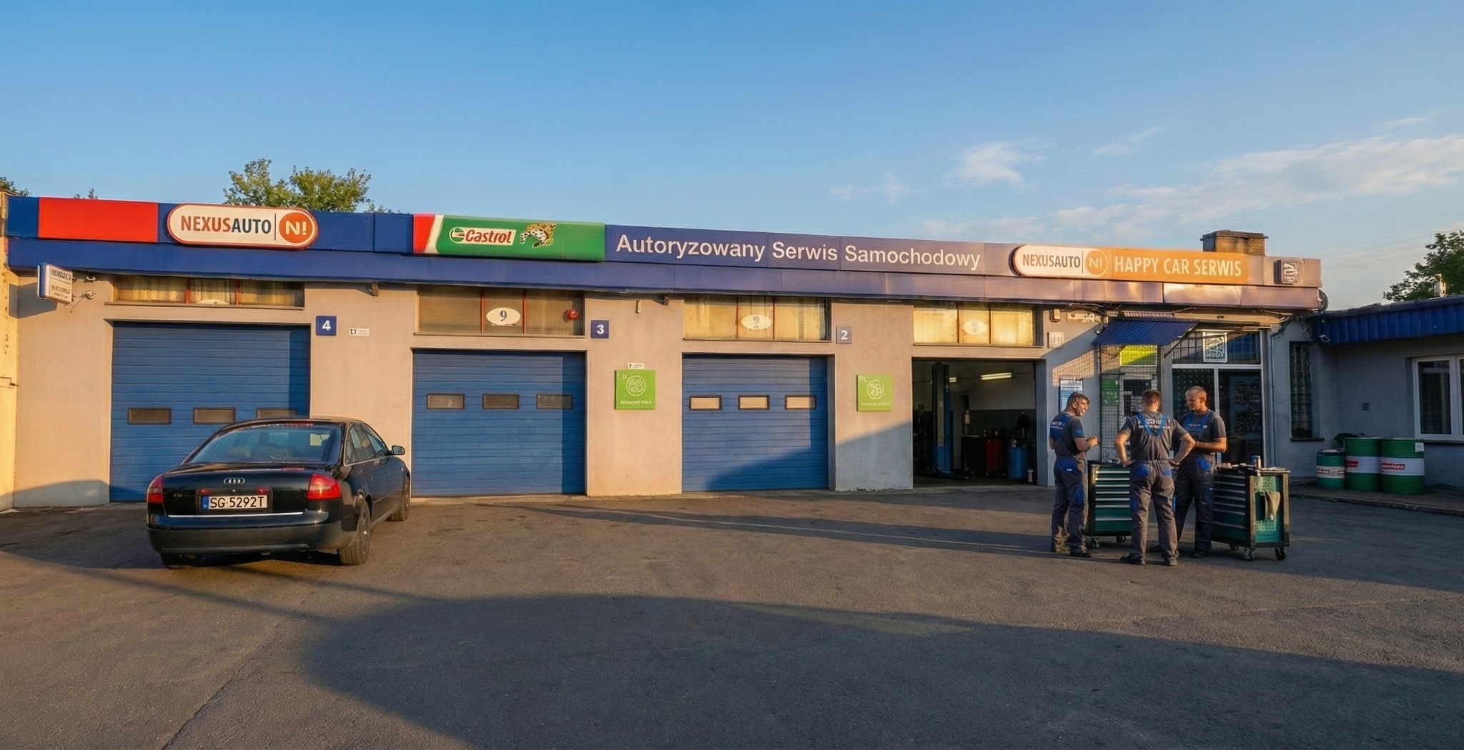 Fasada autoryzowanego serwisu samochodowego z trzema mechanikami w kombinezonach na zewnątrz, zaparkowany samochód Audi A6 i niebieskie bramy garażowe.