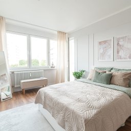 Stalis Staging - Jasna sypialnia z łóżkiem, beżową narzutą w liście, zielonymi poduszkami i minimalistycznymi obrazami na ścianie. Lustro i pufa dopełniają aranżację.