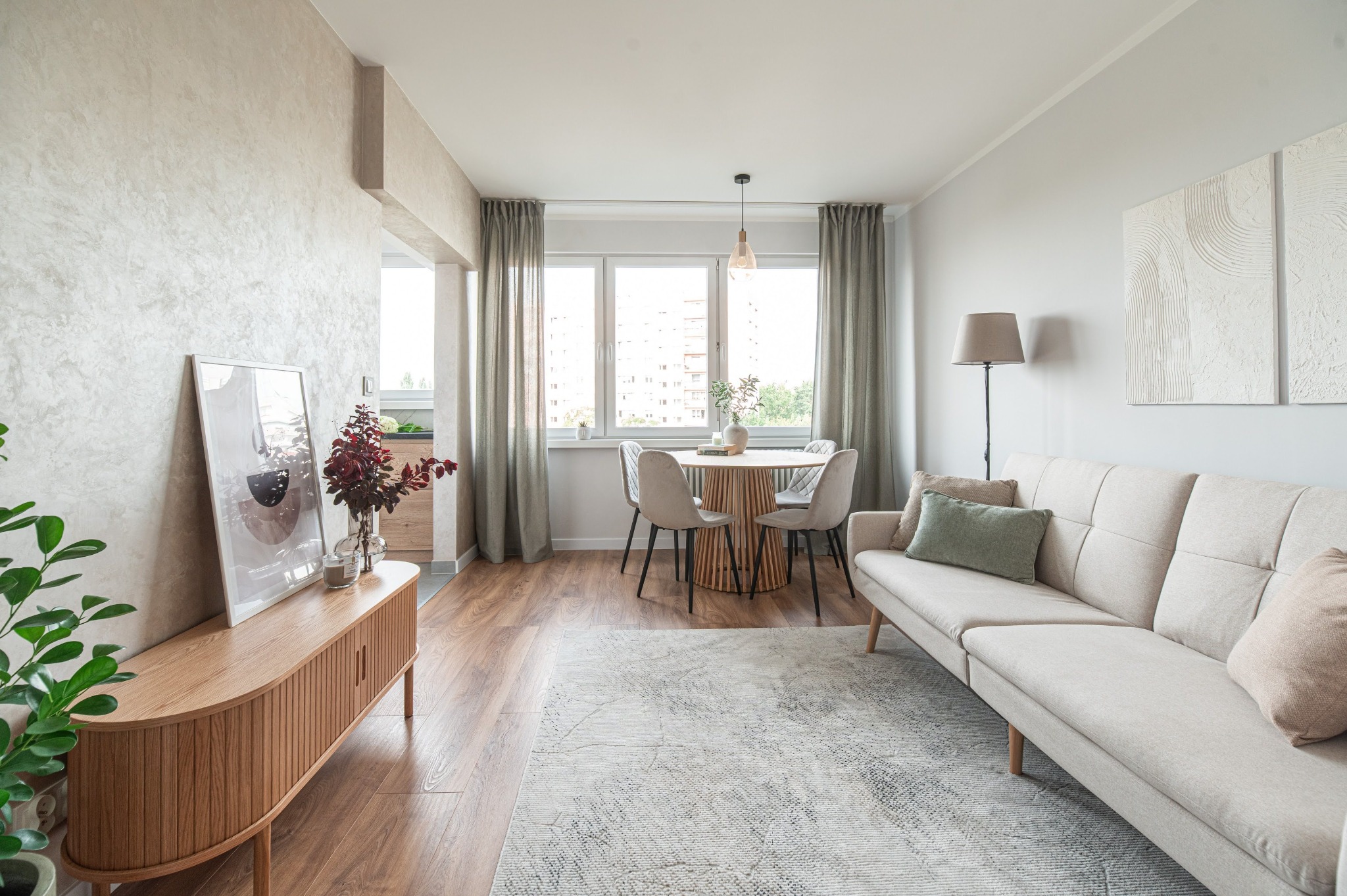 Jasne, przestronne wnętrze mieszkania z drewnianą podłogą, beżową sofą, okrągłym stołem i dekoracjami. Neutralna kolorystyka, minimalistyczny styl.