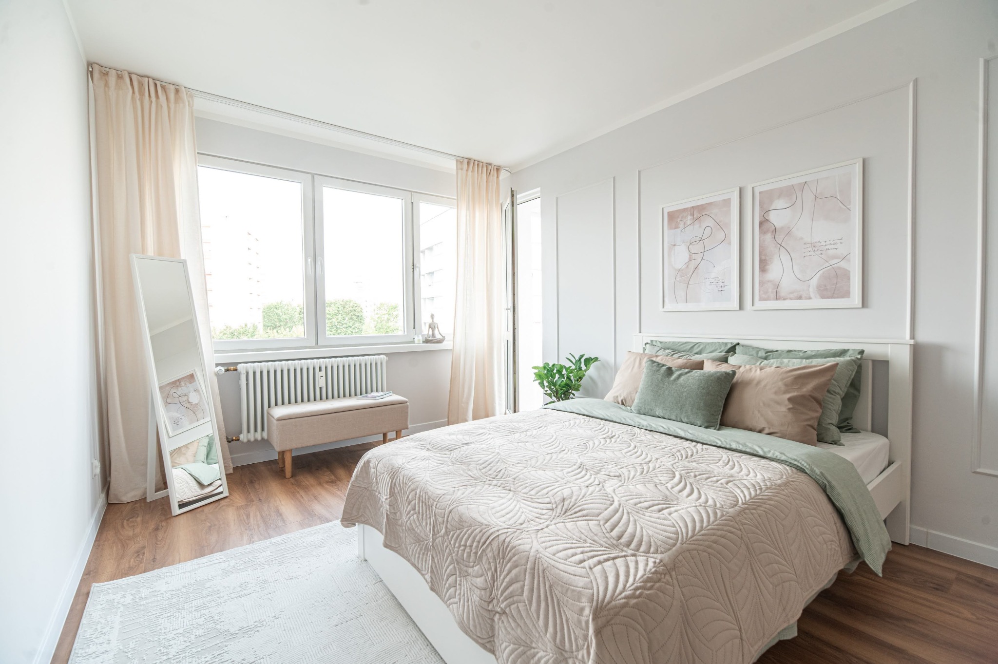 Jasna sypialnia z łóżkiem, beżową narzutą w liście, zielonymi poduszkami i minimalistycznymi obrazami na ścianie. Lustro i pufa dopełniają aranżację.