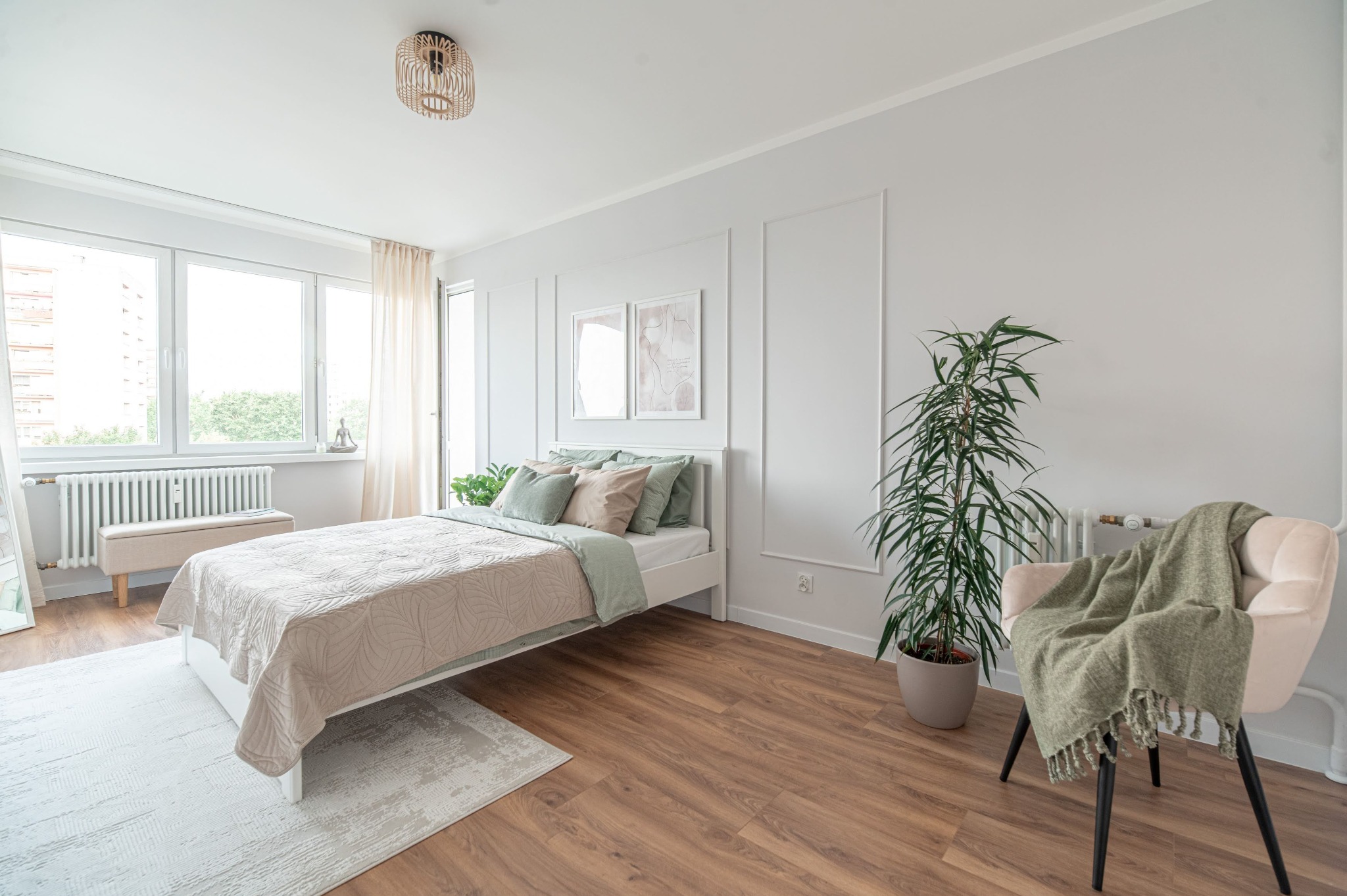 Jasna sypialnia z łóżkiem, rośliną w doniczce i krzesłem z narzutą. Minimalistyczny wystrój, drewniana podłoga, naturalne światło z okna. Dekoracyjne panele ścienne.