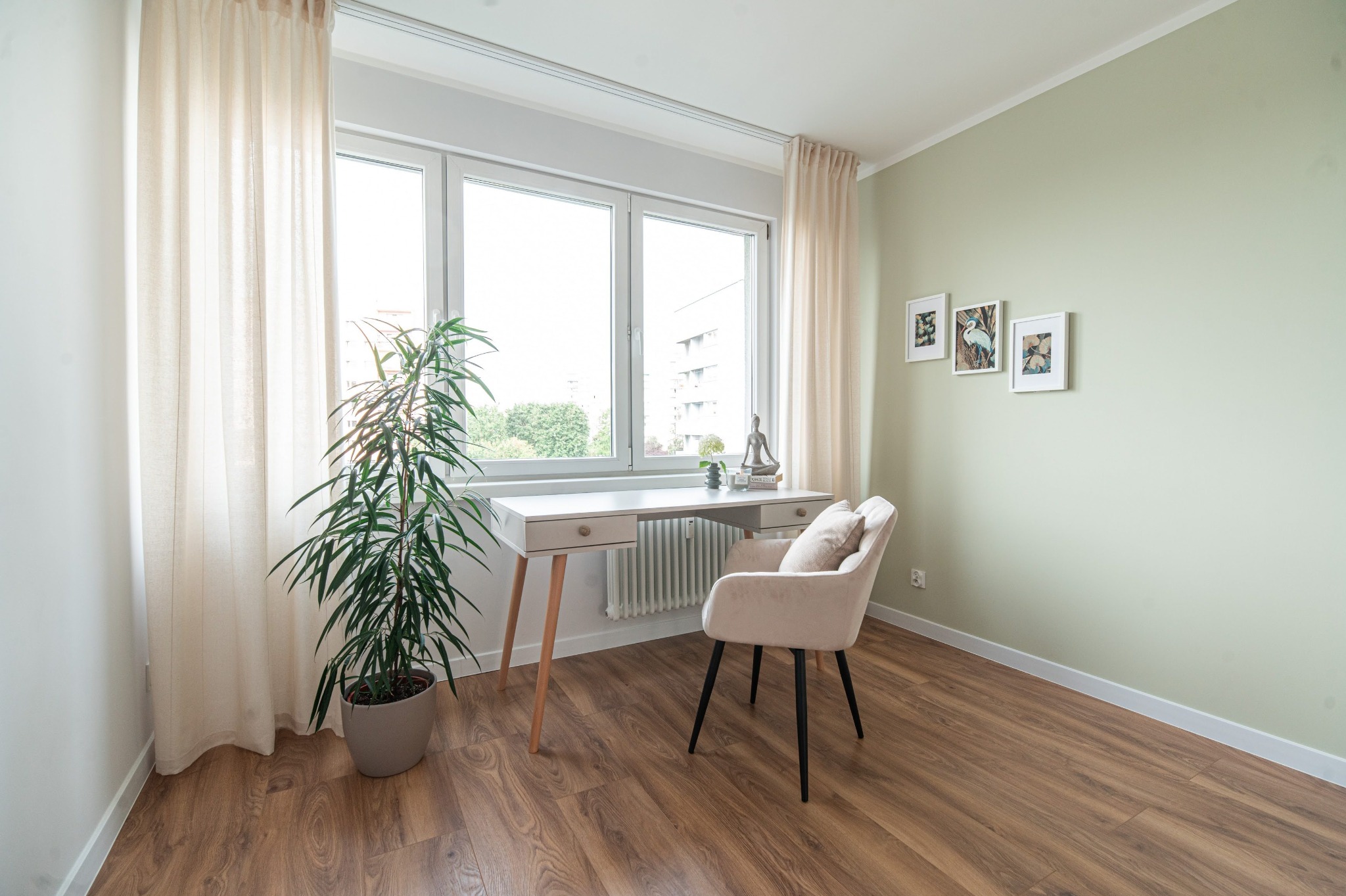 Jasne wnętrze z biurkiem, krzesłem i rośliną doniczkową przy oknie. Na ścianie trzy obrazy w białych ramkach. Podłoga z paneli w odcieniach brązu.