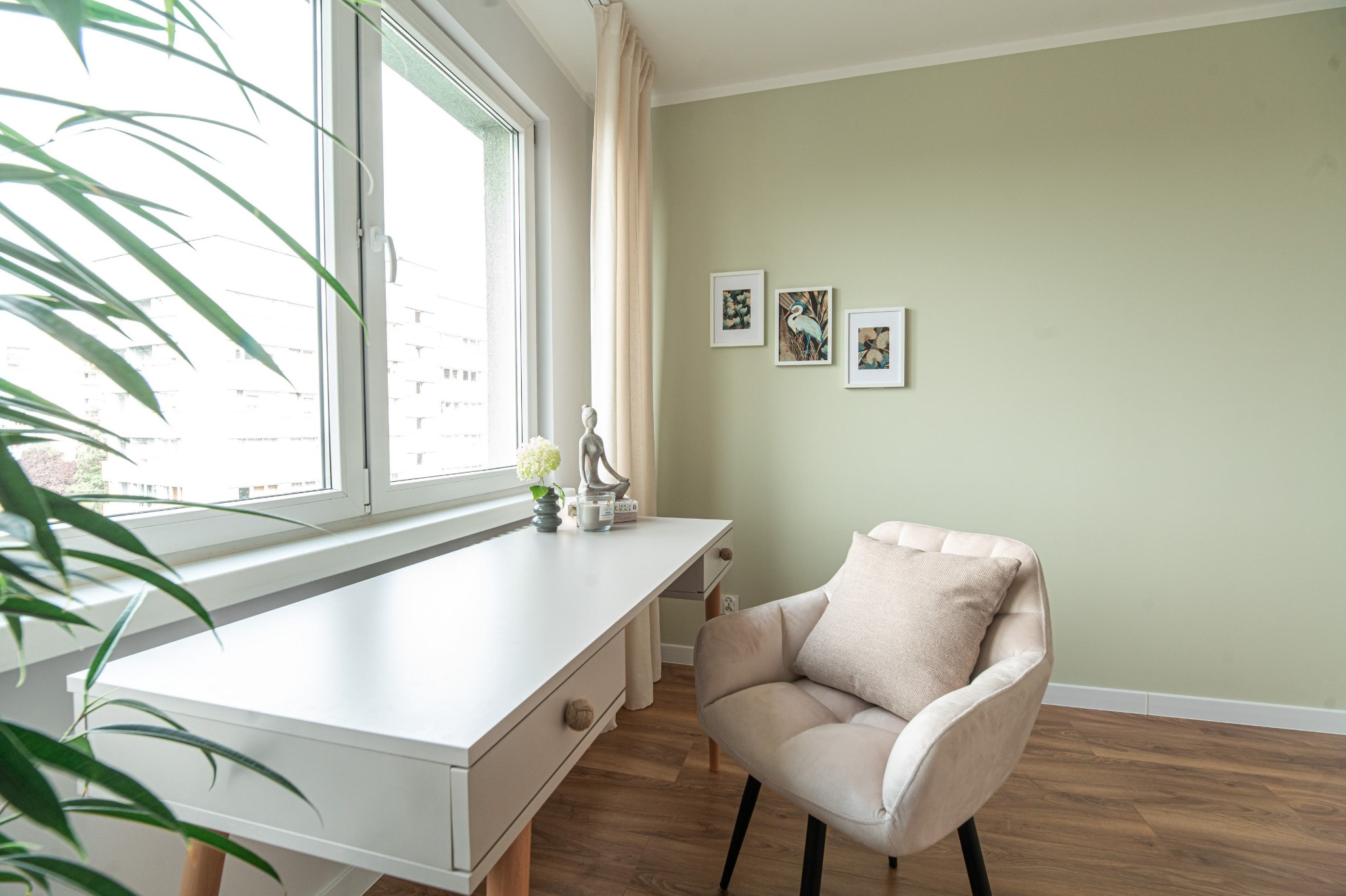 Jasne biuro w mieszkaniu: białe biurko z dekoracjami przy oknie, beżowy fotel z poduszką, zielona ściana z obrazkami. Minimalistyczny, uporządkowany wystrój.