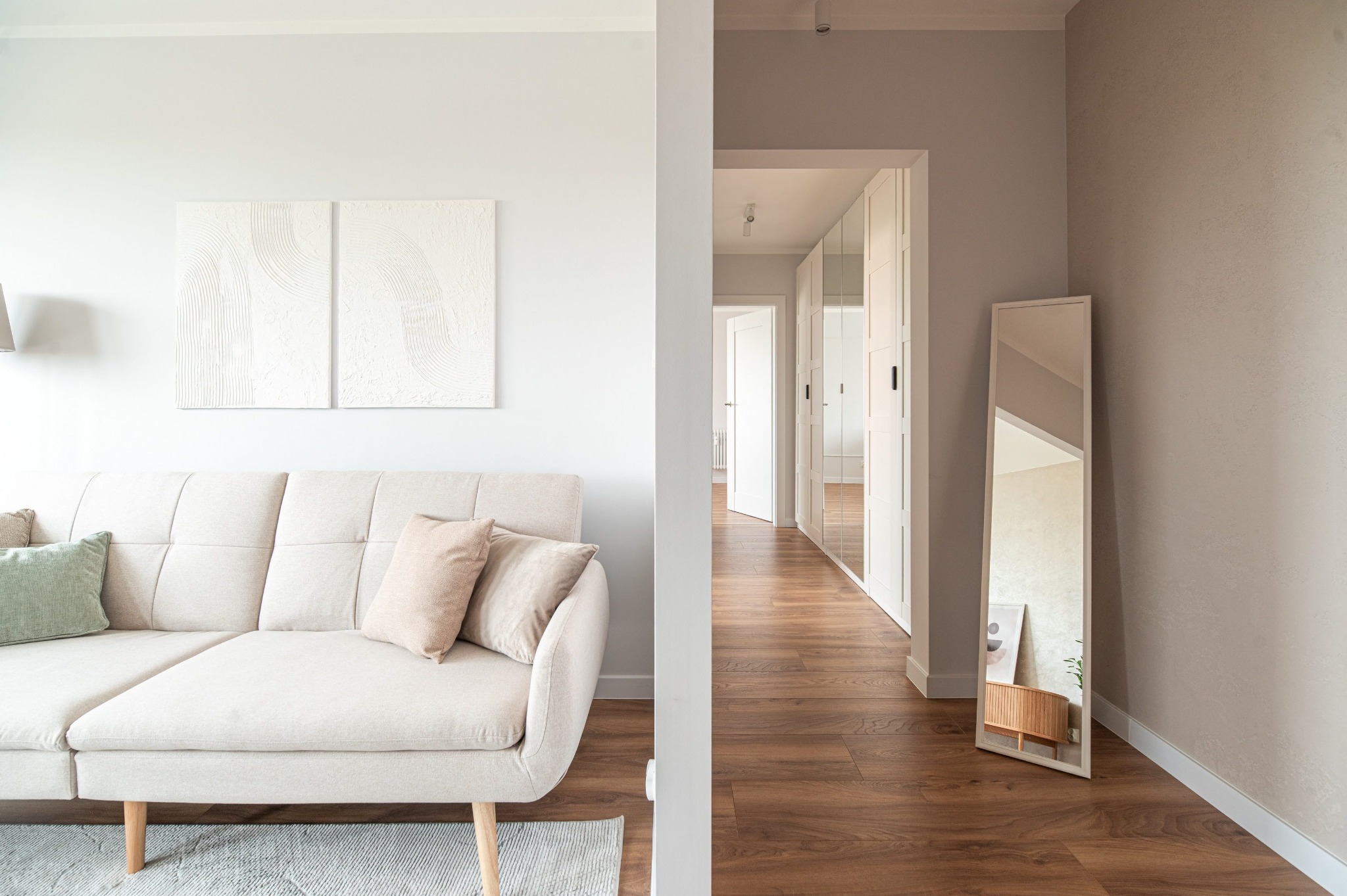 Jasne wnętrze: sofa z poduszkami, abstrakcyjne obrazy na ścianie, lustro i szafa wnękowa w korytarzu. Minimalistyczny design, drewniana podłoga, neutralna kolorystyka.