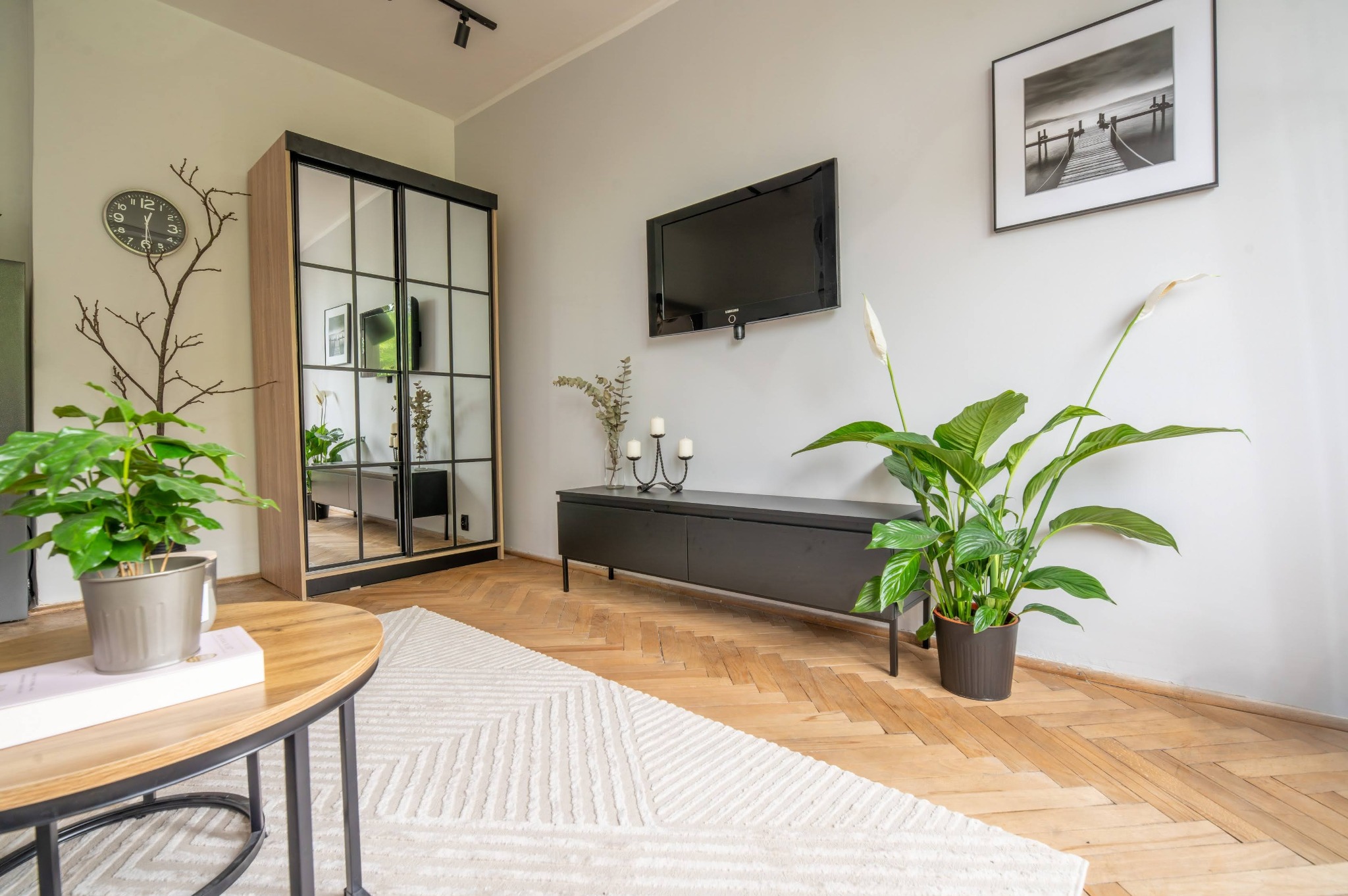 Jasne wnętrze kawalerki z drewnianą podłogą w jodełkę, szafą z lustrami, czarną komodą pod telewizorem i roślinami doniczkowymi. Minimalistyczny wystrój.