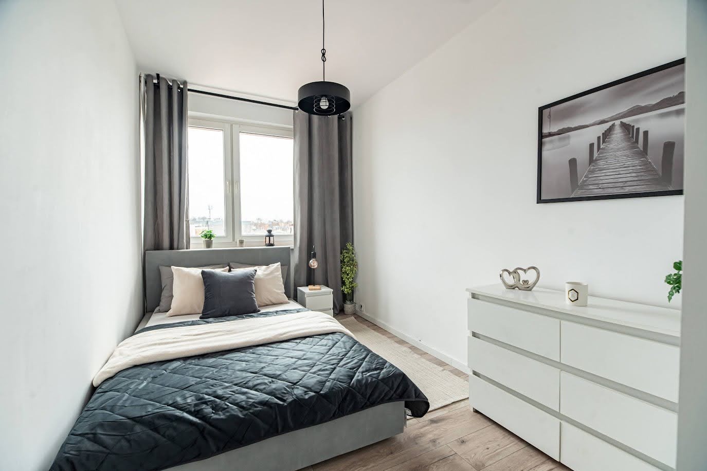 Jasna sypialnia w Szczecinie po liftingu: szare zasłony, łóżko z pikowaną narzutą, biała komoda z dekoracjami, obraz z molo. Przytulne wnętrze gotowe do zamieszkania.