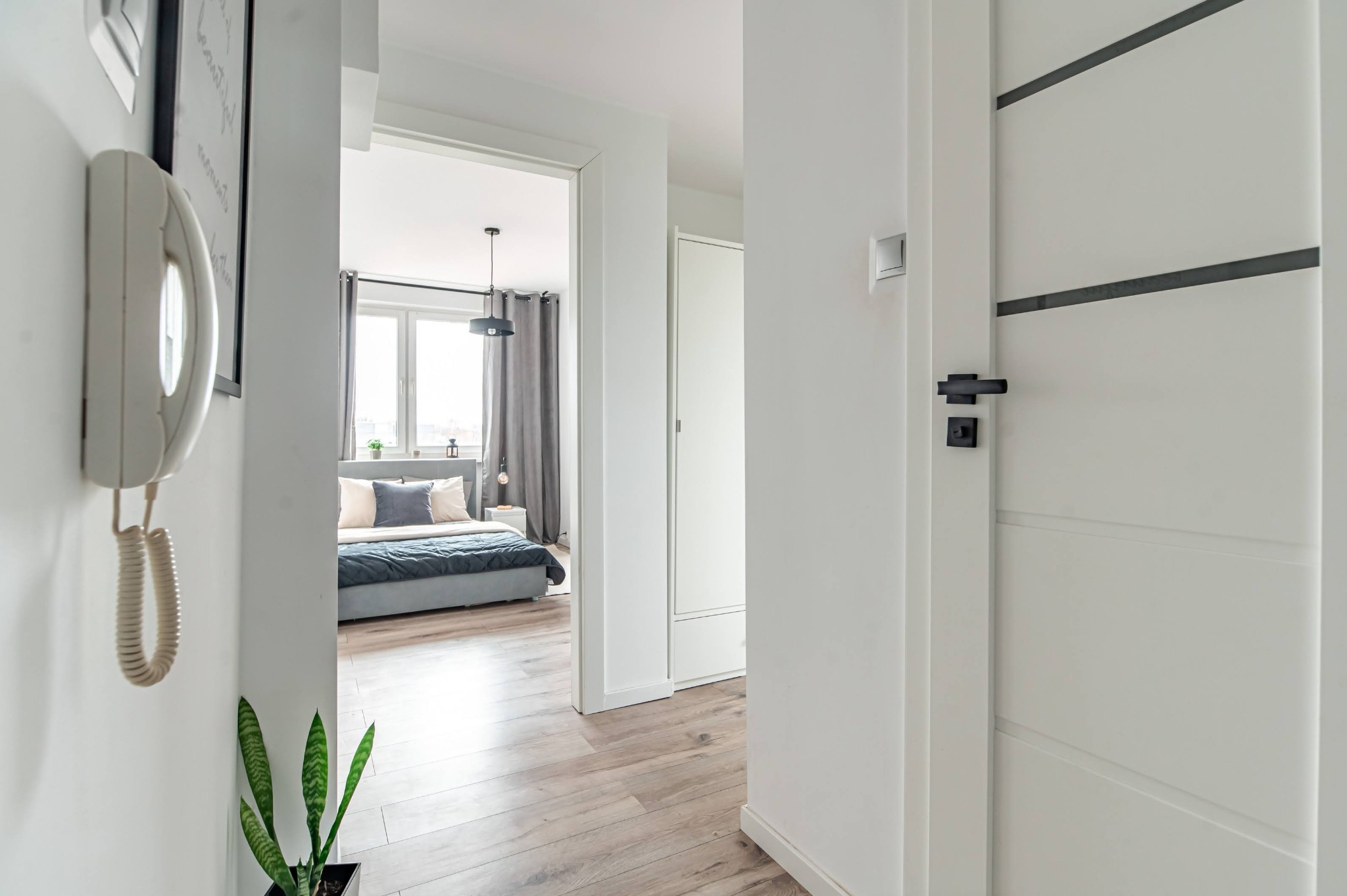 Jasne wnętrze mieszkania z widokiem na sypialnię przez otwarte drzwi. Białe ściany, drewniana podłoga, nowoczesne drzwi z czarnymi akcentami i telefon na ścianie.