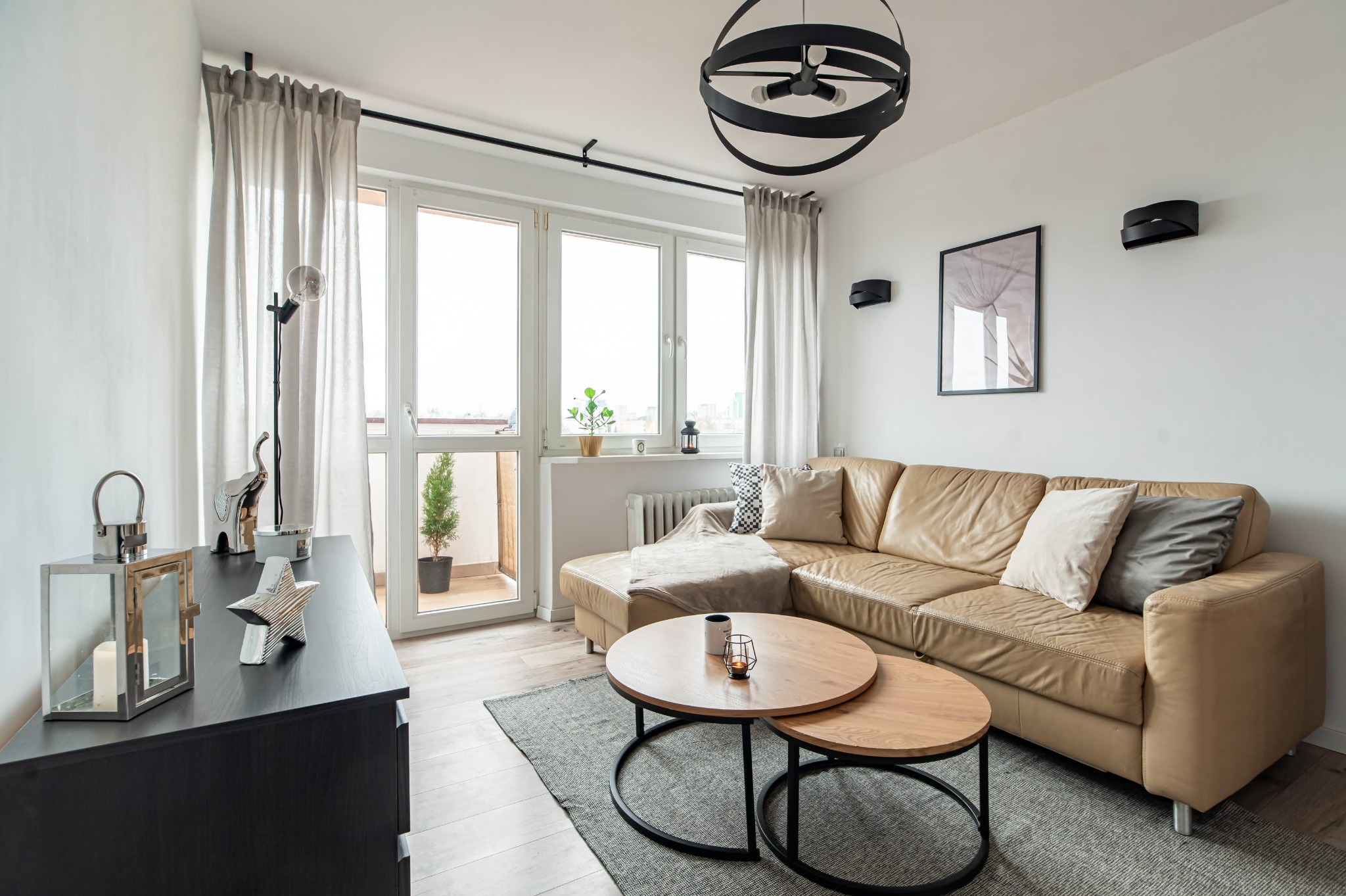 Jasny salon z beżową sofą, okrągłymi stolikami kawowymi i dekoracjami. Widok na balkon z rośliną. Nowoczesne oświetlenie i stonowana kolorystyka.