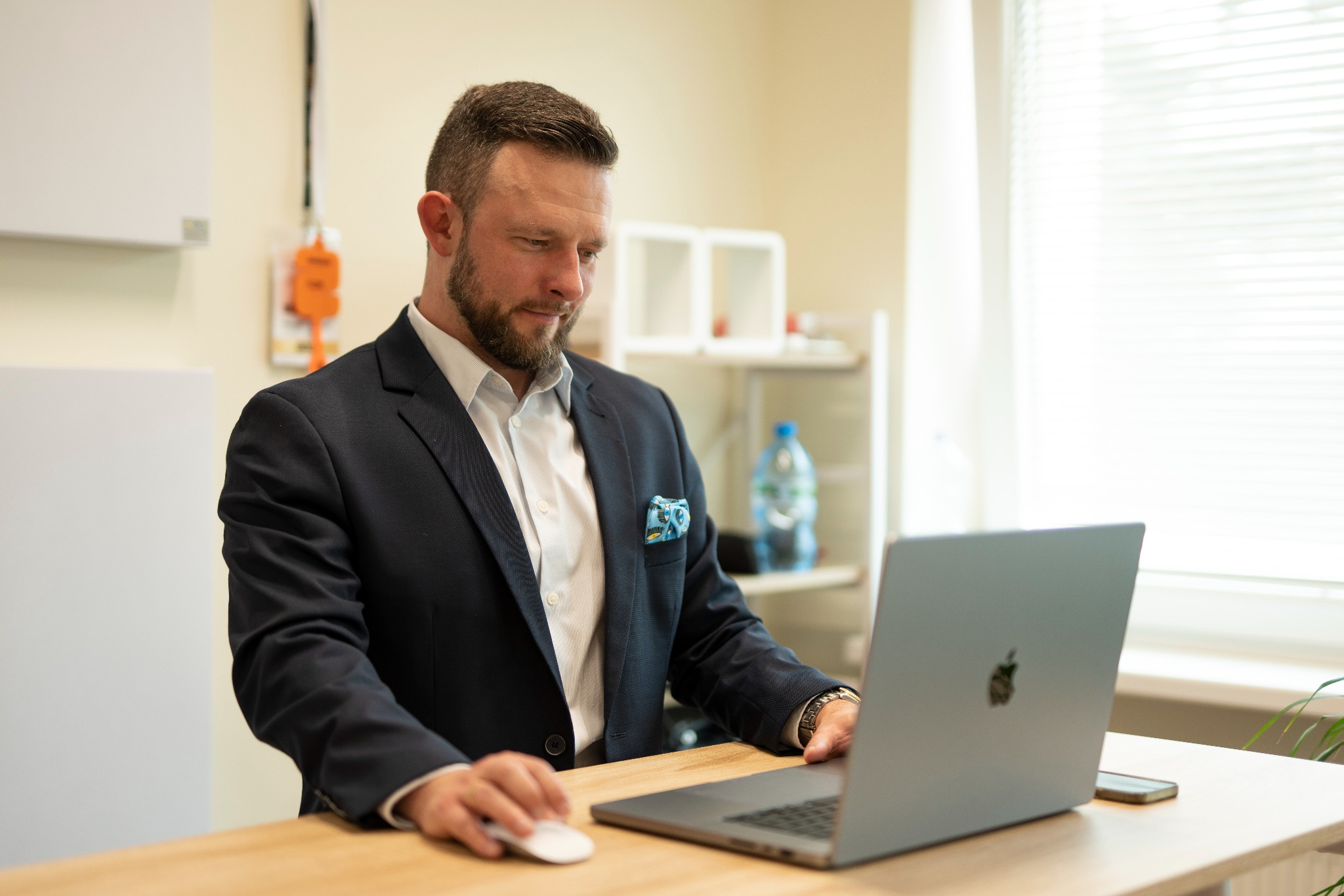 Mężczyzna w garniturze, siedząc przy biurku, pracuje na laptopie. W tle widoczne biurowe wnętrze z oknem i roletami.