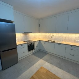 Jan Kaczmarek - Jasna kuchnia z zabudową na wymiar, srebrna lodówka i płytki na ścianie. Ujęcie z niskiego kąta, widoczny fragment podłogi z drewnianym akcentem.