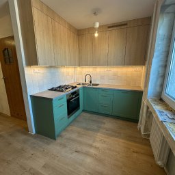 Jan Kaczmarek - Nowoczesna kuchnia z zielonymi szafkami i drewnianymi frontami górnymi, oświetlona listwą LED, z widocznym oknem i grzejnikiem. Ujęcie z perspektywy podłogi.