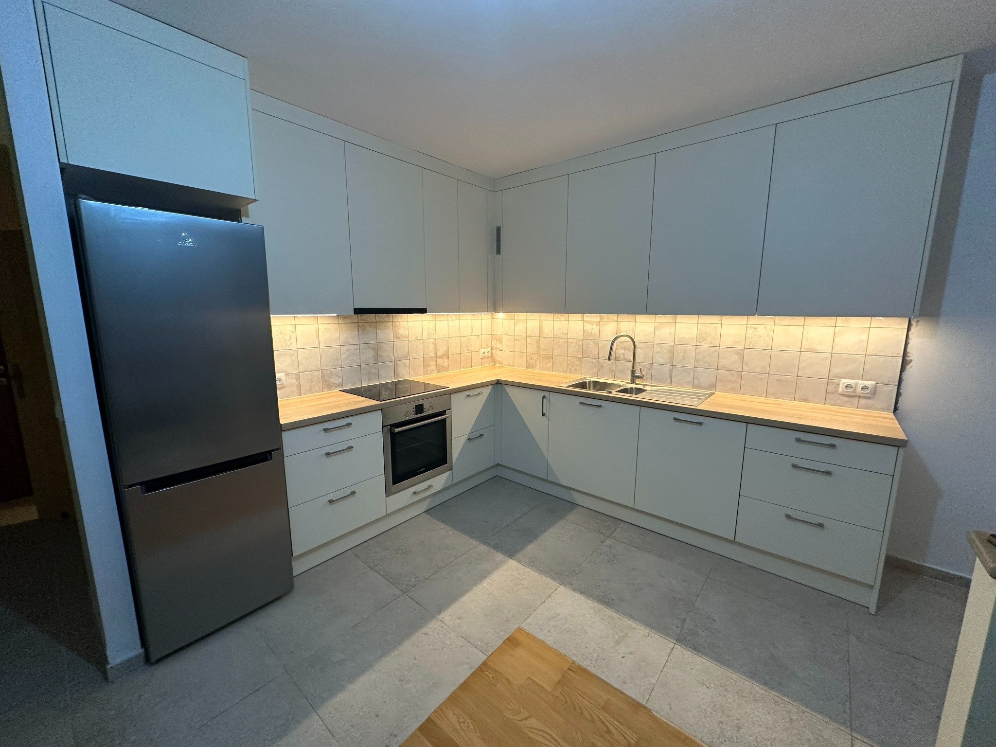 Jasna kuchnia z zabudową na wymiar, srebrna lodówka i płytki na ścianie. Ujęcie z niskiego kąta, widoczny fragment podłogi z drewnianym akcentem.