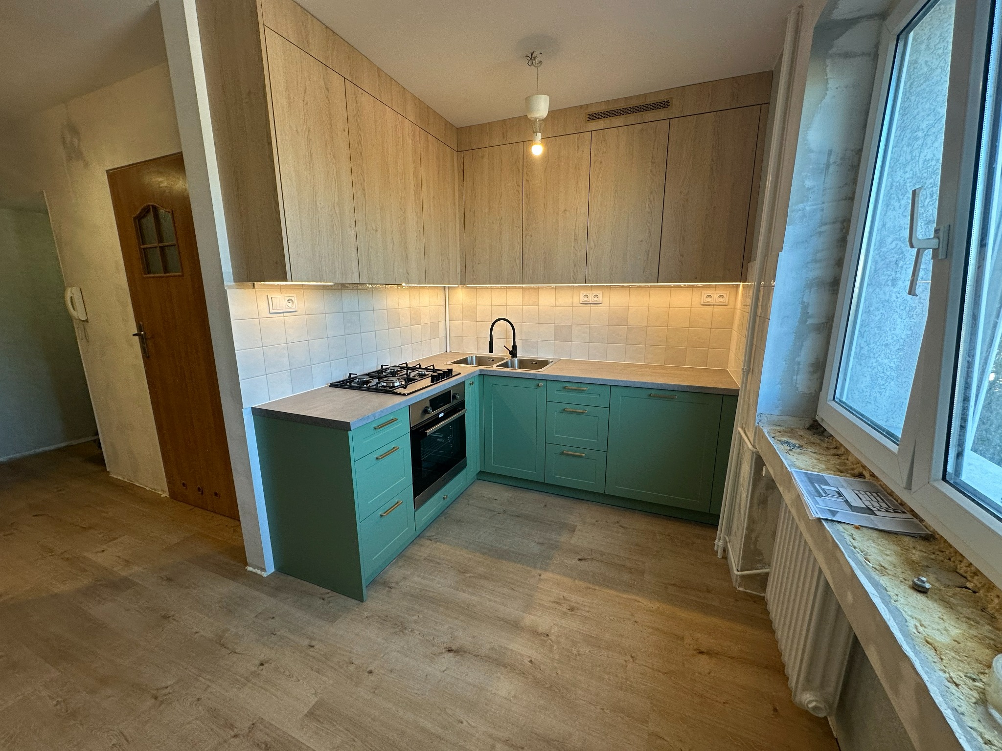 Nowoczesna kuchnia z zielonymi szafkami i drewnianymi frontami górnymi, oświetlona listwą LED, z widocznym oknem i grzejnikiem. Ujęcie z perspektywy podłogi.