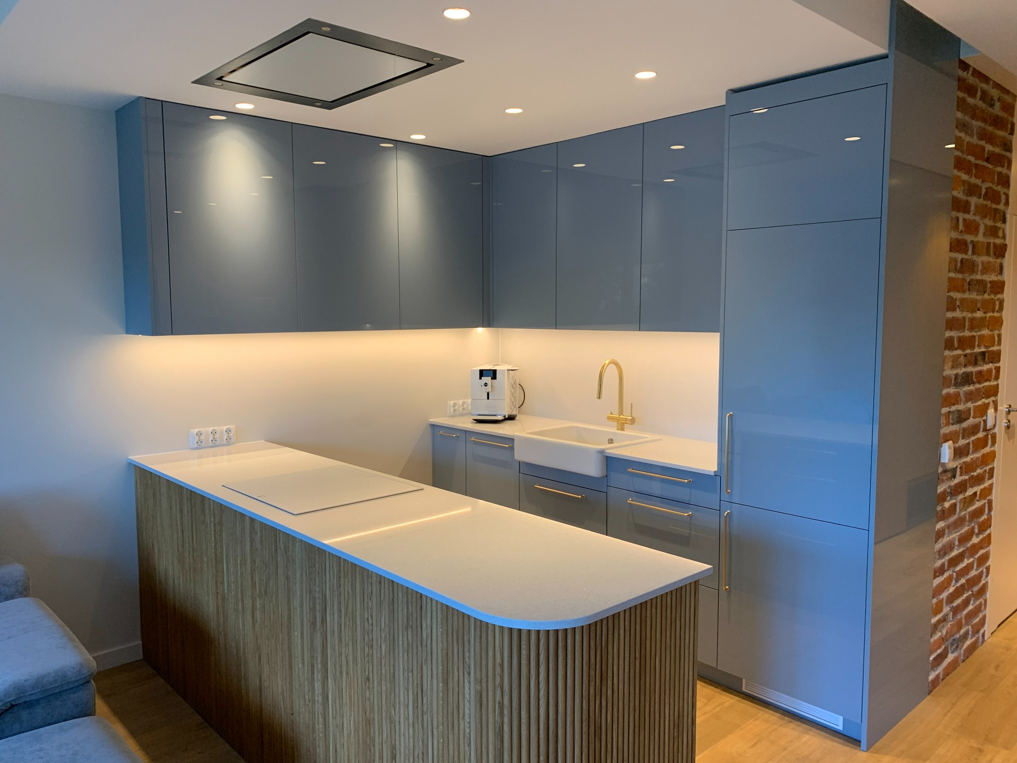 Niebieska kuchnia na wymiar z drewnianą wyspą i złotą armaturą, oświetlona punktowo, z fragmentem ściany z cegły w tle. Nowoczesny design.