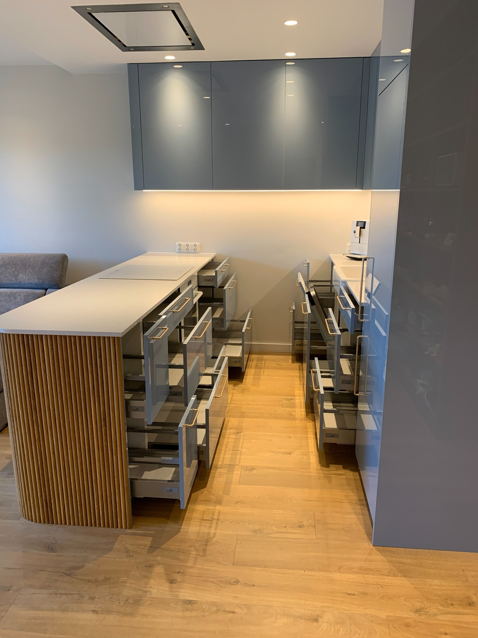 Nowoczesna kuchnia z szarymi, lakierowanymi frontami i drewnianym wykończeniem wyspy. Otwarta przestrzeń z wysuwanymi szufladami i oświetleniem podszafkowym.