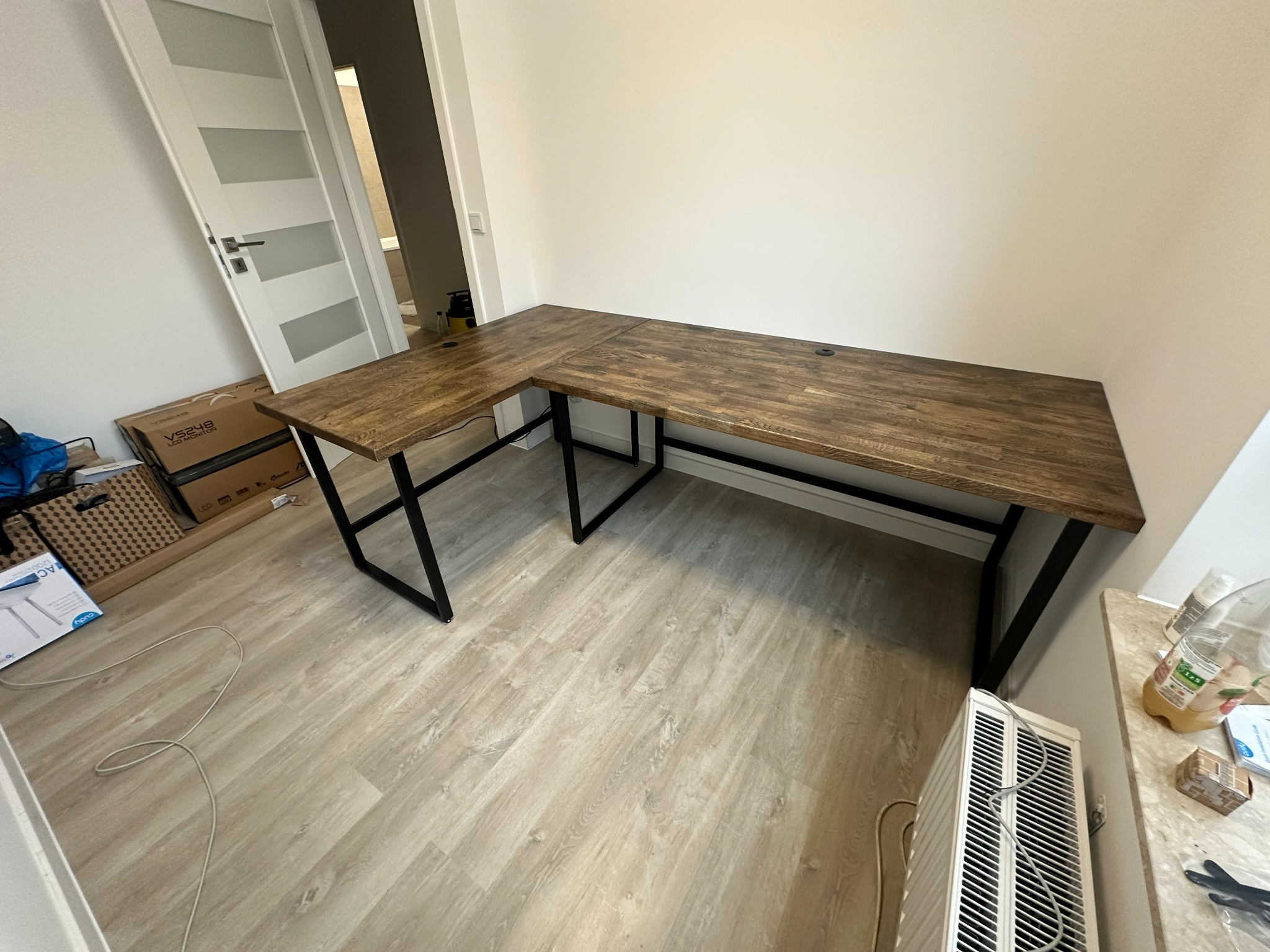 Drewniane biurko narożne z czarnymi nogami w jasnym wnętrzu. Widoczne gniazda na kable. Na podłodze panele, przy ścianie białe drzwi i grzejnik.
