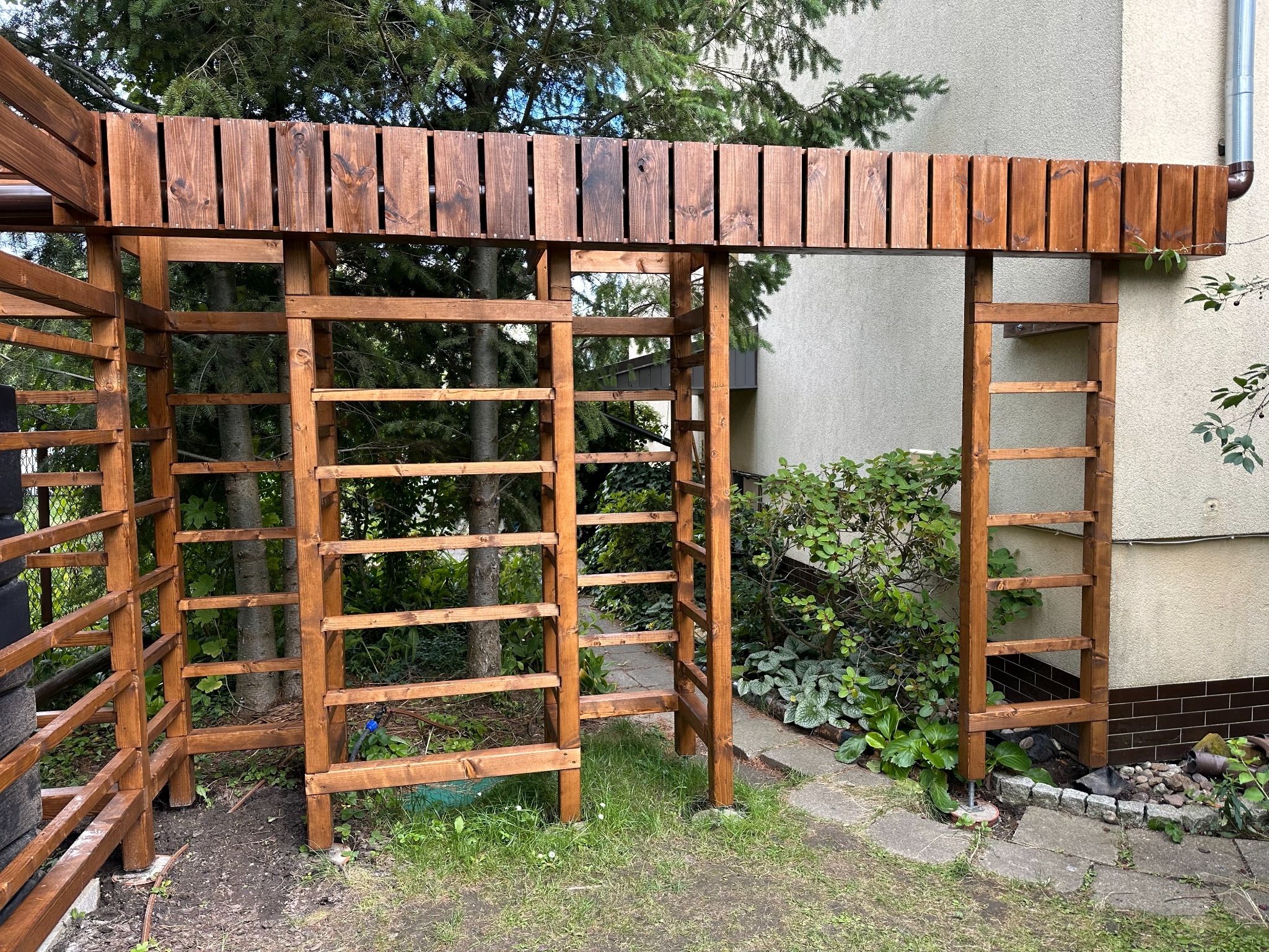 Drewniana pergola w ogrodzie, z widoczną konstrukcją z belek i desek, pomalowana na brązowo, z zielenią w tle i fragmentem elewacji budynku.