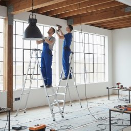 ZEUS Taras HERZHII - Dwóch monterów na drabinach instaluje dużą, czarną lampę w przestronnym, jasnym pomieszczeniu z drewnianym stropem i dużymi oknami. Widoczne narzędzia i zabezpieczenia podłogi.