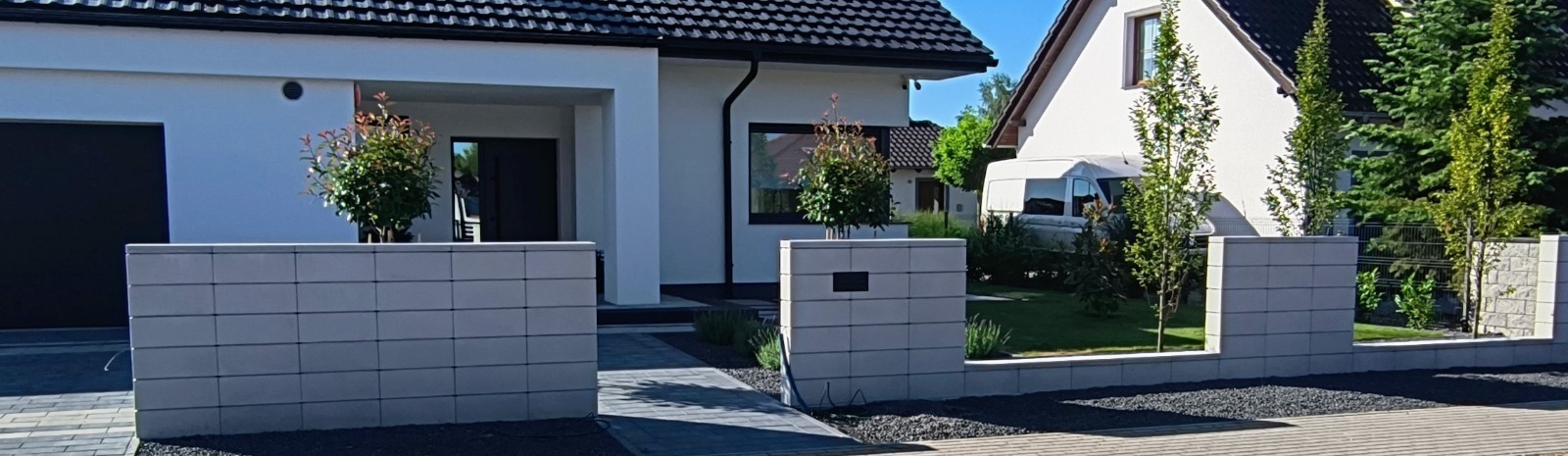 Nowoczesny ogród frontowy z kostki brukowej i niskim murem z bloczków, ozdobiony drzewkami w donicach. Widoczny dom z czarnym dachem i elementami wykończenia.