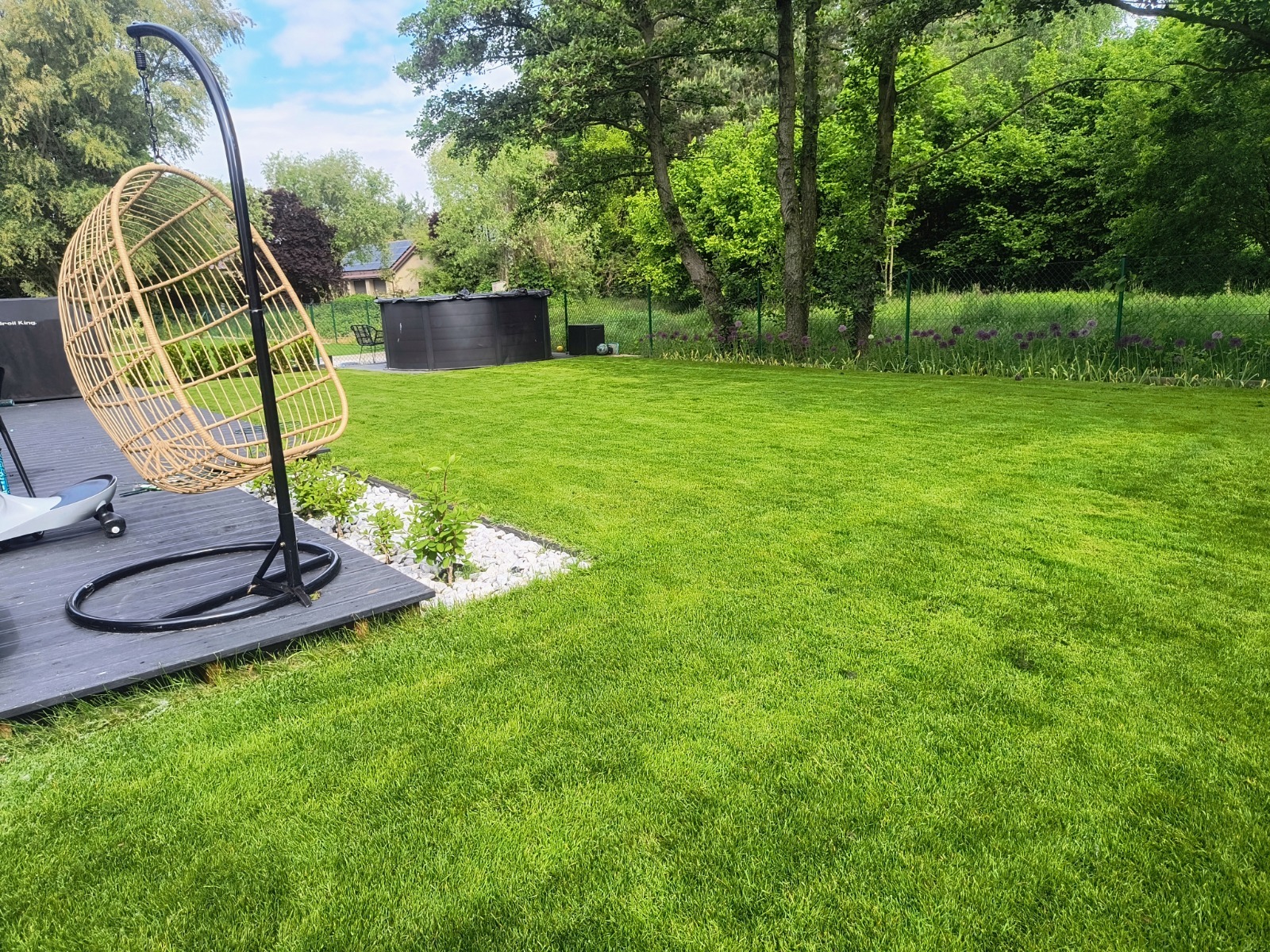 Zadbany trawnik z równo przystrzyżoną trawą, graniczący z drewnianym tarasem i dekoracyjnym żwirem. W tle widoczny basen ogrodowy i drzewa.